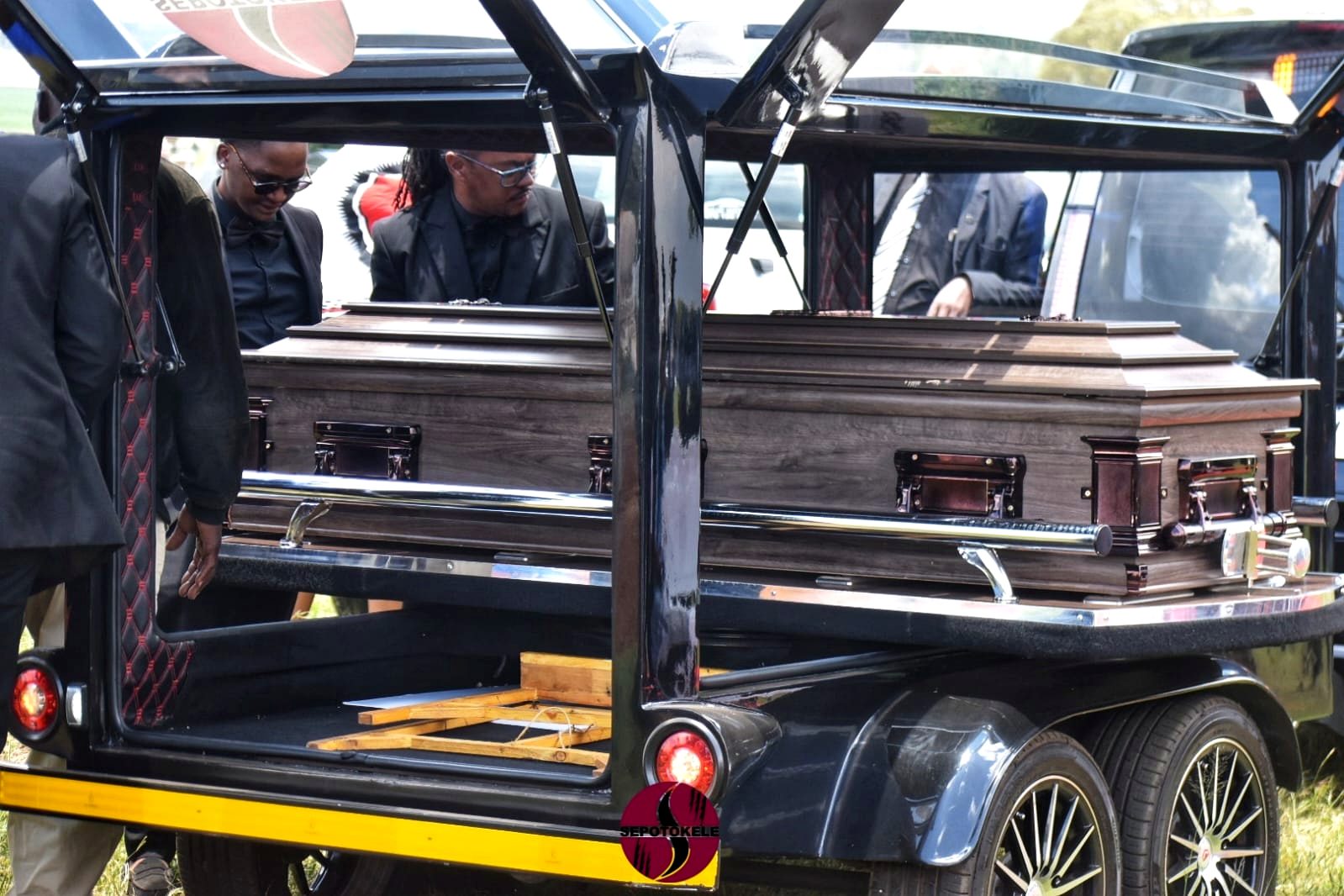 Pallbearers in black suits at the open Sepotokele glass hearse trailer